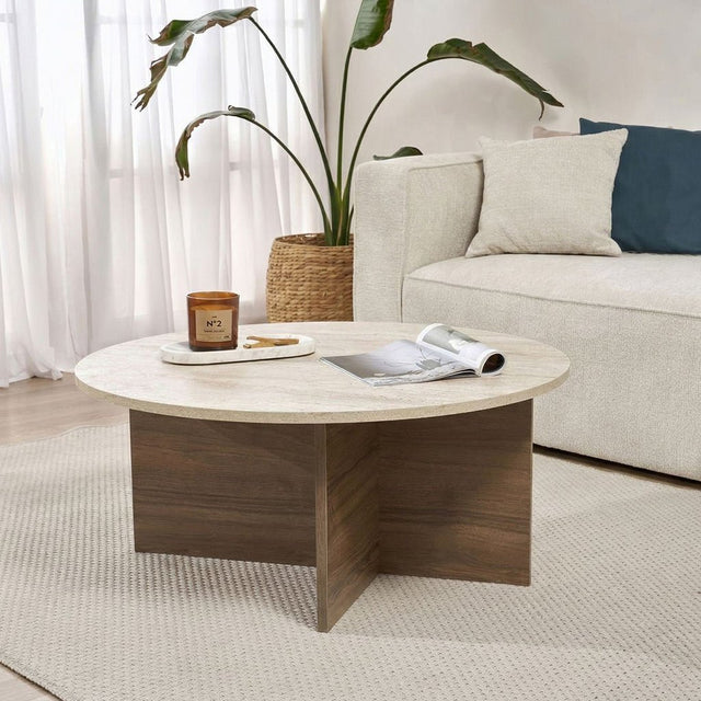 Ronde salontafel met travertin blad en donker houten onderstel in kruisvorm. Op de achtergrond bank en plant.